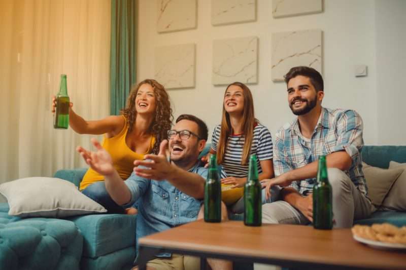 Grupo de amigos jóvenes que se divierten viendo la televisión y bebiendo cervezas en casa en una vida compartida.