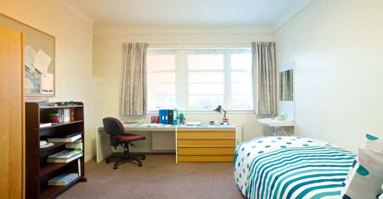 Habitación de estudiante linda en un campus universitario con una cama individual, un escritorio grande, un armario y una gran ventana.