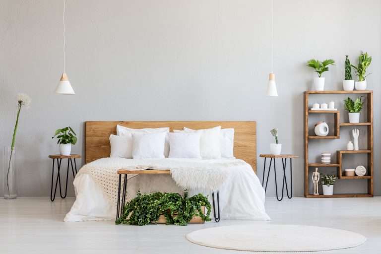 Minimal interior de una habitación amueblada con una cama doble de madera con almohadas blancas y plantas, una alfombra de pelo blanco y muebles modernos de madera rodeando la cama.