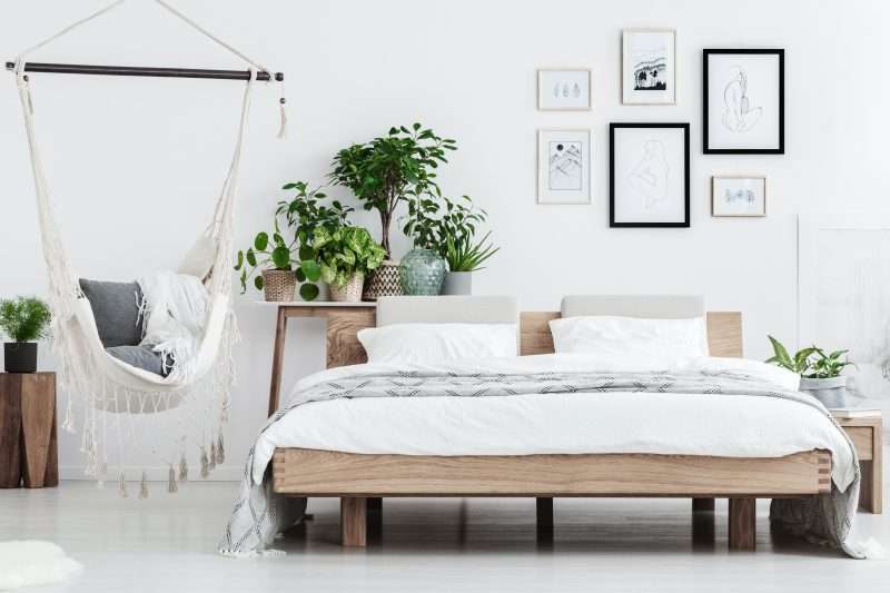 Plantas detrás de una cama de madera cerca de una hamaca con almohadas en un interior de habitación natural con carteles en la pared blanca