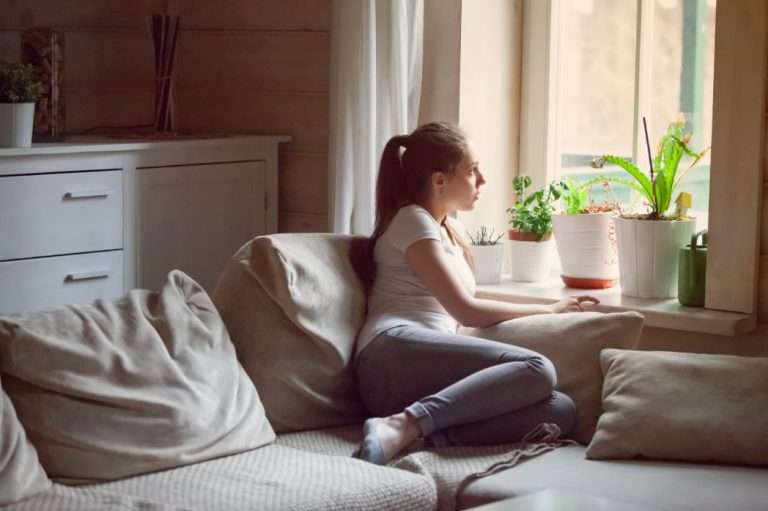 Mujer milenaria atractiva sentada sola en un sofá en la sala de estar de su casa pensando mirarando por la ventana. Parece relajada, triste y melancólica.