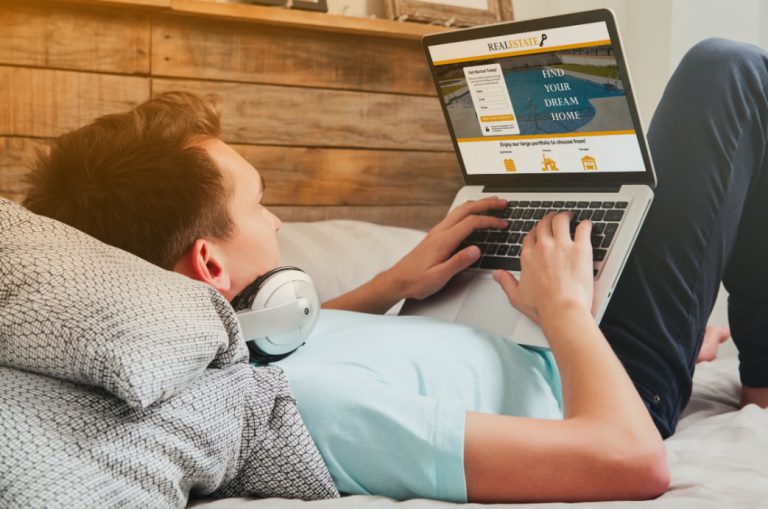 Hombre jóven caucásico acostado en su cama en casa buscando una habitación en una casa en un sitio web de viviendas con un computador