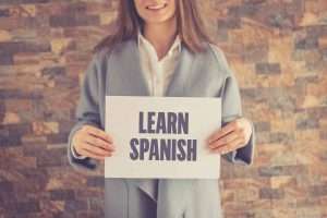 Caucasian girl standing in front of a brick wall holding a sign saying "Learn Spanish"