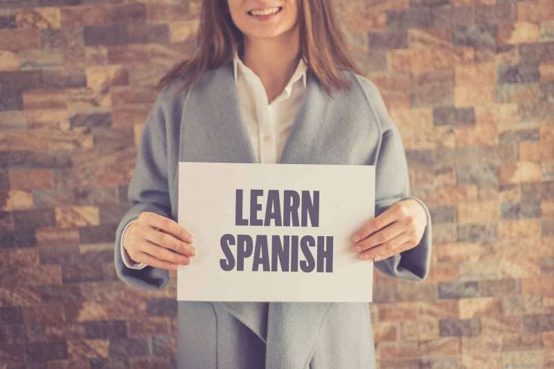 Caucasian girl standing in front of a brick wall holding a sign saying "Learn Spanish"