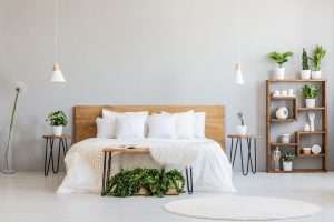 Minimal bedroom interior furnished with a double wooden bed with white pillows and plants, a white furry rug and modern wooden furniture surrounding the bed