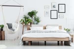 Plants behind wooden bed near hammock with pillows in natural bedroom interior with posters on white wall
